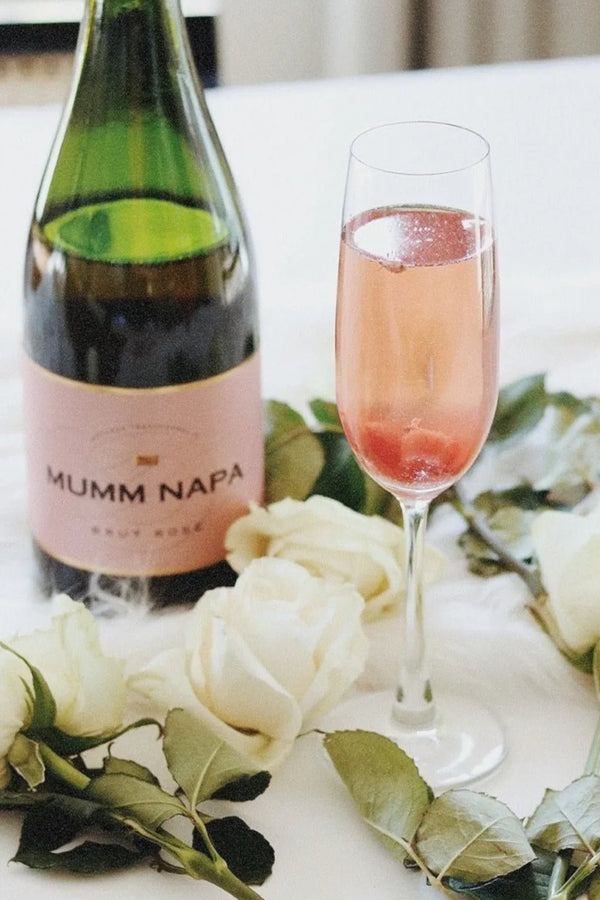 A bottle of Mumm Napa Brut Rosé wine next to a glass of pink sparkling wine, surrounded by white roses and greenery.