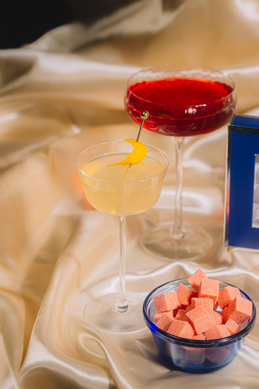 Two cocktails on a satin surface with a small bowl of pink cubes.