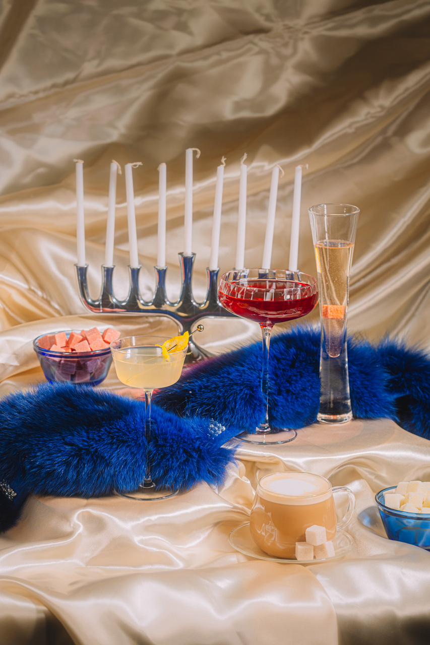 Decorative table setting with candles, glasses of drinks, and a teapot on a draped fabric background.