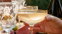 A gold-rimmed coupe glass filled with a sparkling botanical cocktail, held during an elegant outdoor Damp January social gathering