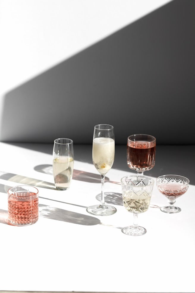 A collection of six various glass drinks, including champagne and other colorful beverages, displayed on a white surface with shadows.