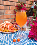Glass of Aperol spritz with a bottle of sparkling wine, plate of sliced oranges, and flowers on a blue mosaic table.