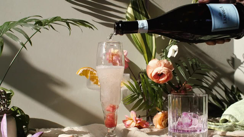 A glass is being filled with a sparkling drink from a bottle, surrounded by flowers and greenery.