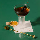 A stylish cocktail in a coupe glass garnished with olives and an orange slice, resting on a napkin with a gold spoon beside it, against a green backdrop.