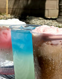 Three colorful drinks in clear cups on a table: a pink layered drink with whipped cream, a blue icy drink, and a brown fizzy drink topped with pink foam.