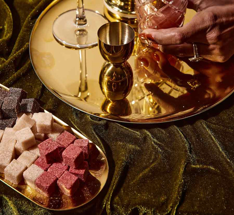A gold tray with assorted colored sugar cubes (white, pink, and dark) and a cocktail glass being held by a hand, set against a green velvet background.