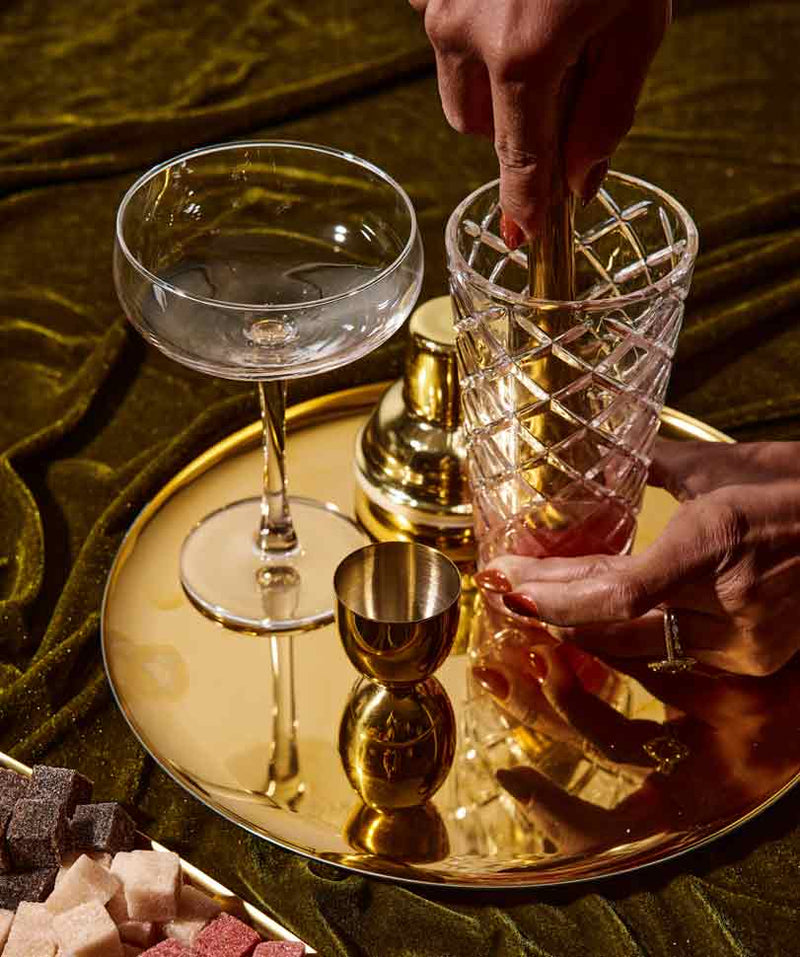 A hand is muddling ingredients in a crystal glass with a golden tool, next to a coupe glass and a jigger on a shiny gold tray, surrounded by colorful sugar cubes.