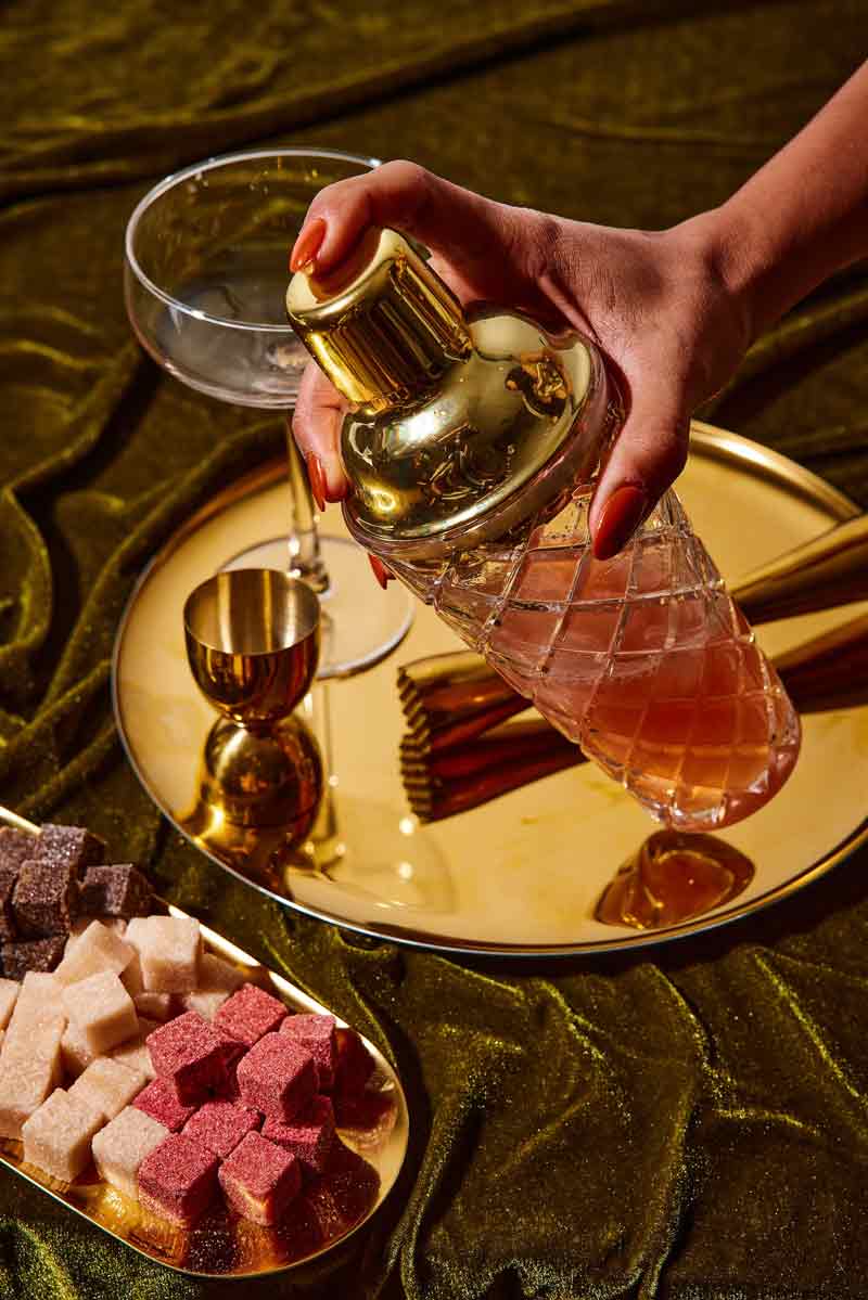 A hand holding a gold cocktail shaker above a gold tray with a martini glass, measuring cup, and colorful sugar cubes on a green velvet surface.