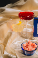 Two cocktails on a satin surface with a small bowl of pink cubes.