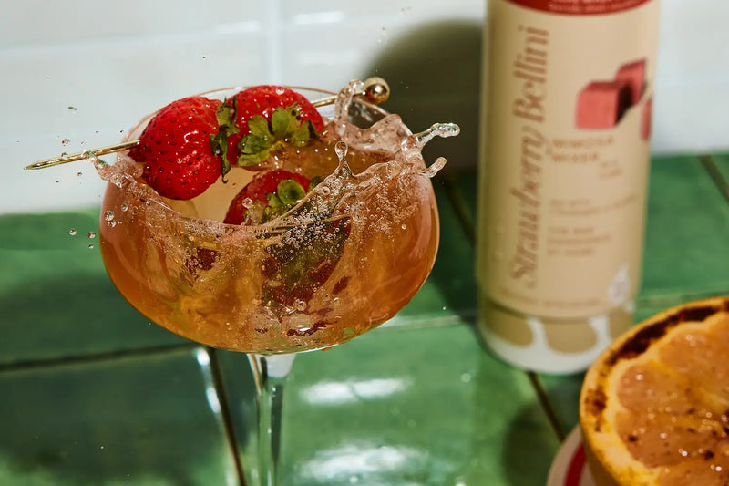 A cocktail glass filled with a sparkling drink, fresh strawberries garnish, and droplets of liquid splashing around. A bottle labeled "Strawberry Bellini" is visible in the background.
